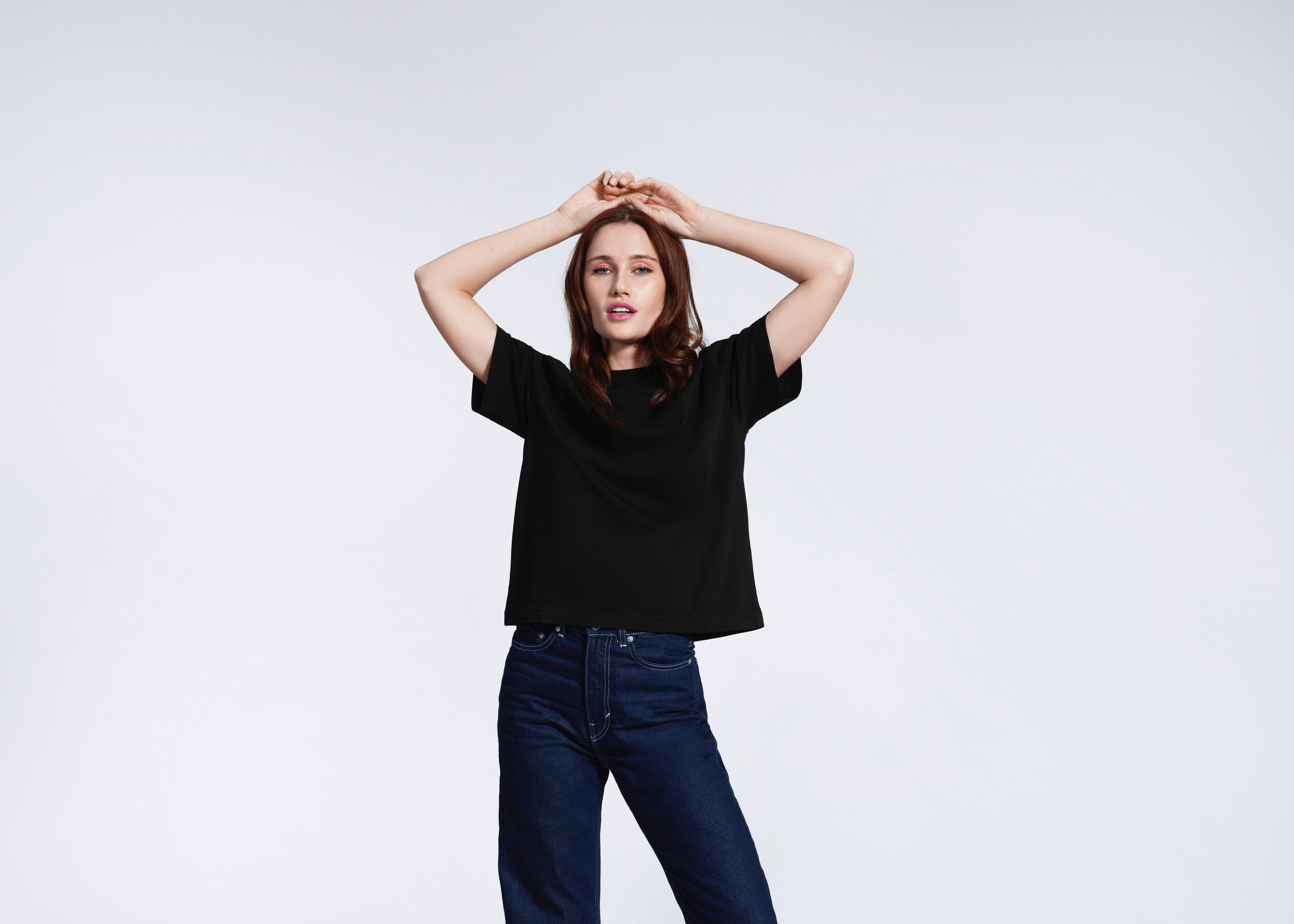 A woman with her hands above her head looking at the camera, modelling a black boxy t-shirt.