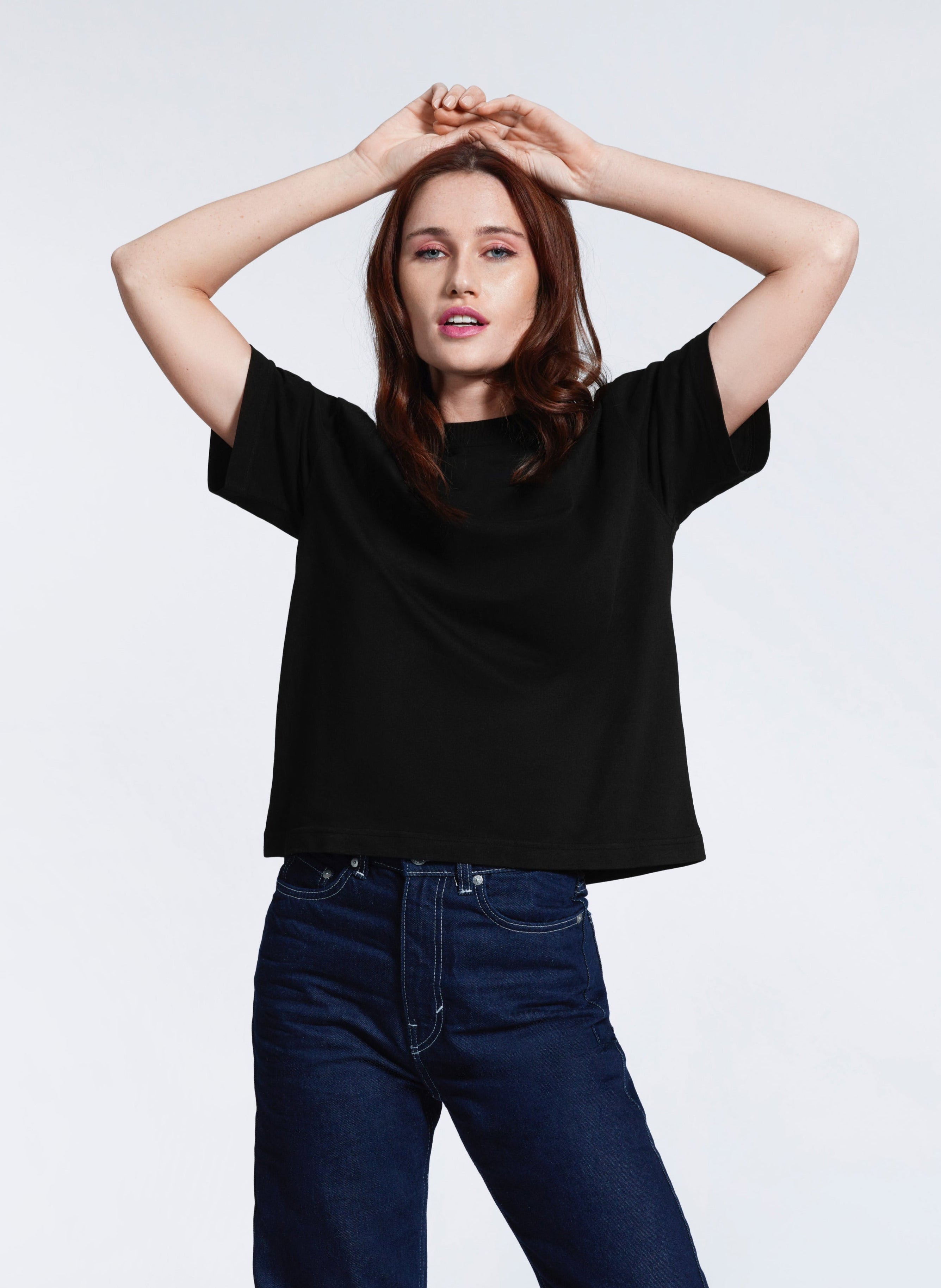 A woman with her hands above her head looking at the camera, modelling a black boxy t-shirt.